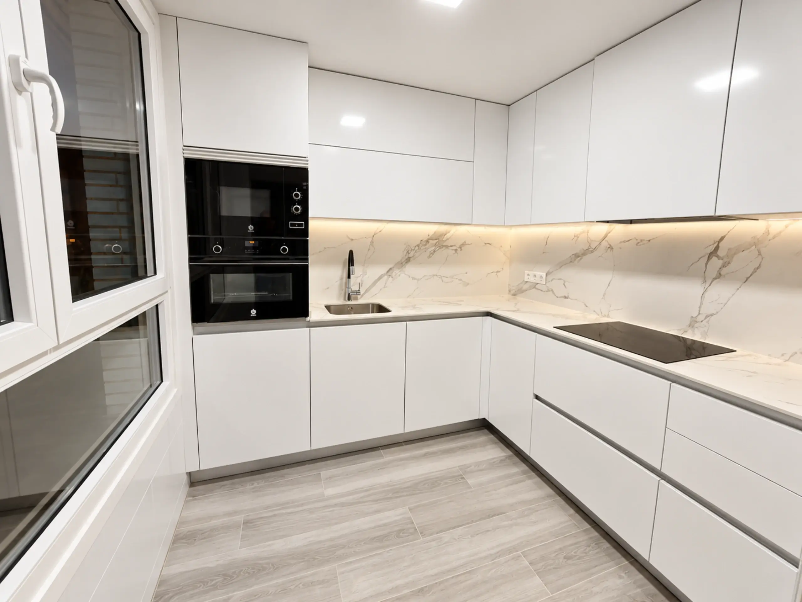 Cocina en L blanca con luz LED bajo los muebles altos y frontal de efecto mármol